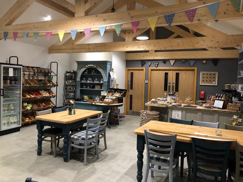 Interior of a cozy café or small shop with wooden tables and chairs, exposed wooden beams on the ceiling, colorful triangular pennant bunting hanging across, shelves with fresh produce and packaged goods, and a counter displaying baked items and beverages.