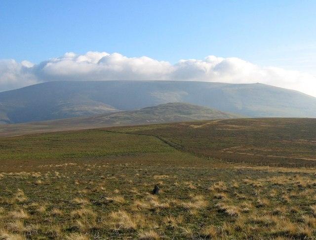 Rolling grassy landscape with gentle hills in the foreground, leading up to a larger, cloud-capped mountain in the background.