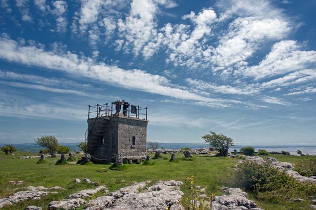 Stone building with a tower situated in a grassy area with scattered rocks, under a sky with scattered clouds.