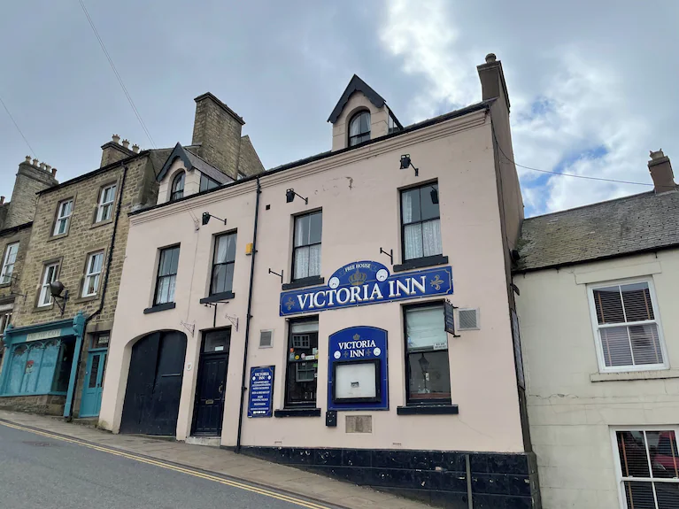 Three-storey building on a street corner with a sign reading 'Victoria Inn' above the entrance, featuring cream walls with black trim and adjacent stone buildings under a cloudy sky
