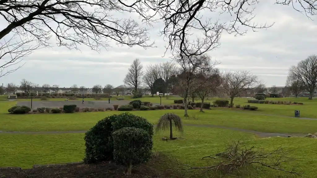 Bare trees and green lawns in Vulcan Park, Workington, with a paved basketball court visible in the distance under a cloudy, overcast sky.