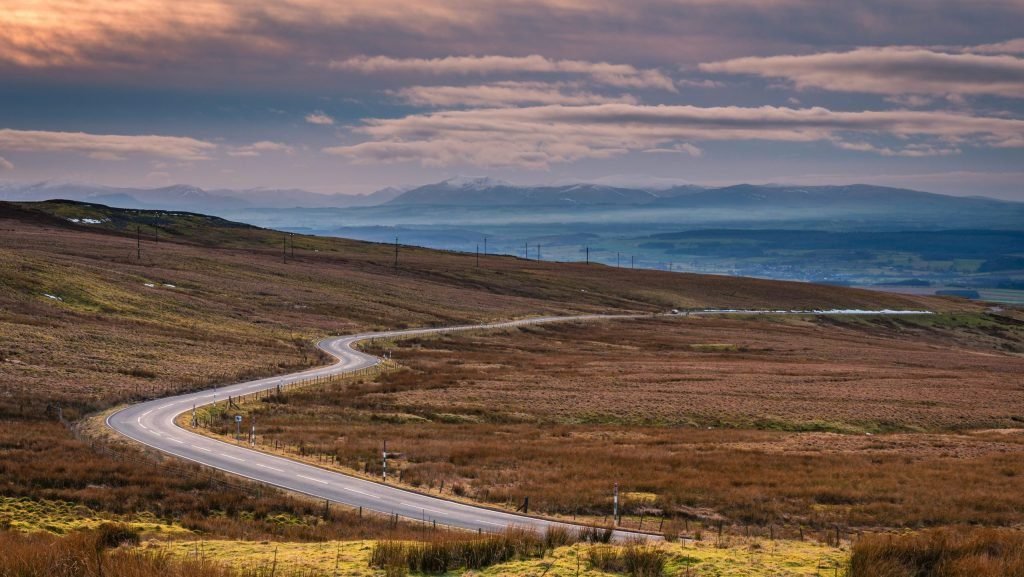 Winding road through a vast, brownish-orange moorland with distant mountains under a cloudy sky at dusk.