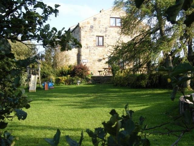 Stone house with wooden window frames surrounded by a green lawn and leafy trees, with children’s slide and play equipment to the left and a wooden picnic table near the back of the house