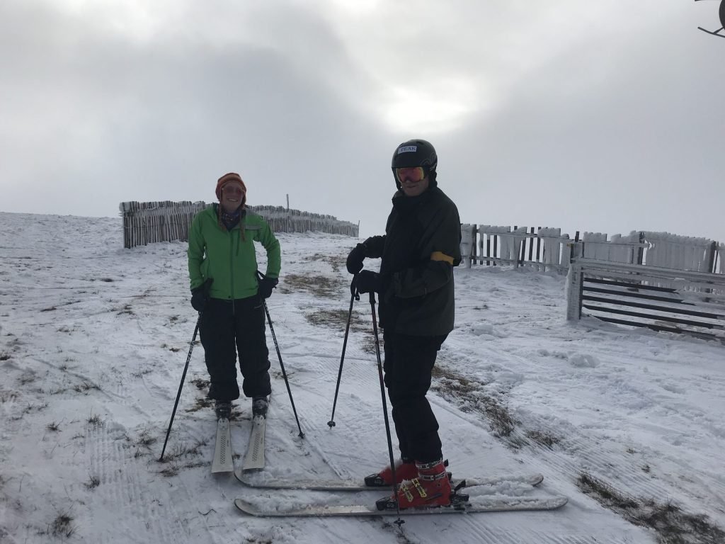 Helvellyn | Guardian of Glenridding and Patterdale 7 Two skiers standing on snow near wooden snow fences under a cloudy sky, one wearing a green jacket and orange hat, the other in a black helmet and dark clothing with red ski boots.