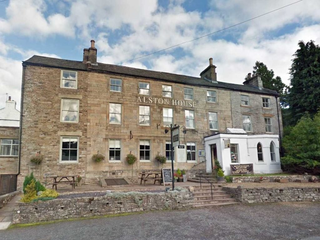 Large three-story stone building with six windows on each floor and a white entrance extension, labelled Alston House, with picnic benches outside and a stone stairway leading to the entrance under a partly cloudy sky