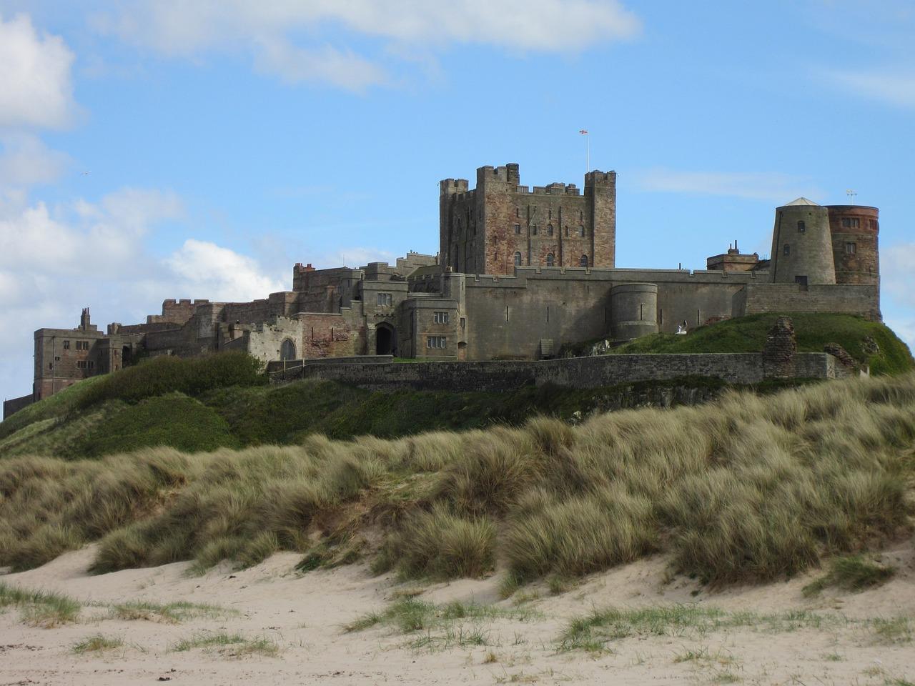 A historic castle situated on a grassy hill, with sand dunes in the foreground and a blue sky with clouds in the background.