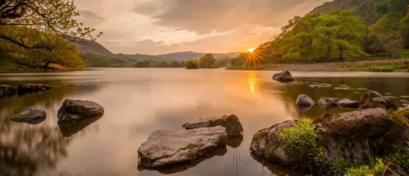 Calm lake at sunset with rocks in the foreground, surrounded by green trees and hills.