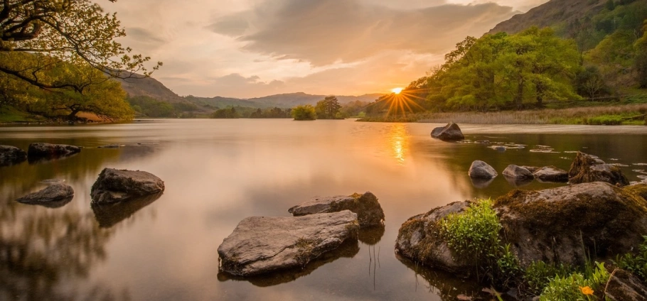 Calm lake at sunset with rocks in the foreground, surrounded by green trees and hills.