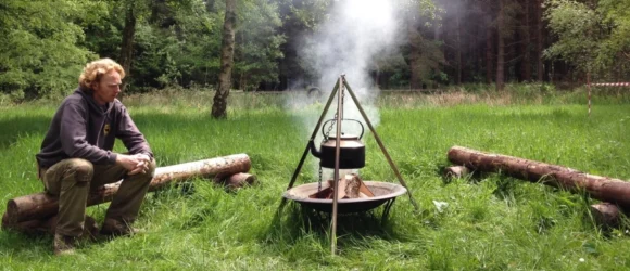 A person sitting on a log beside a campfire with a kettle hanging over it, in a grassy clearing surrounded by trees.