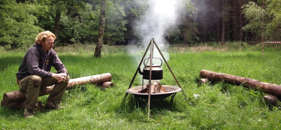 A person sitting on a log beside a campfire with a kettle hanging over it, in a grassy clearing surrounded by trees.