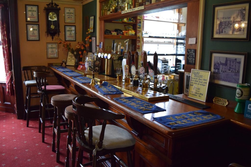 Interior of a traditional pub with a wooden bar counter, stools, several beer taps, framed pictures and certificates on the walls, and blue bar mats arranged on the counter.
