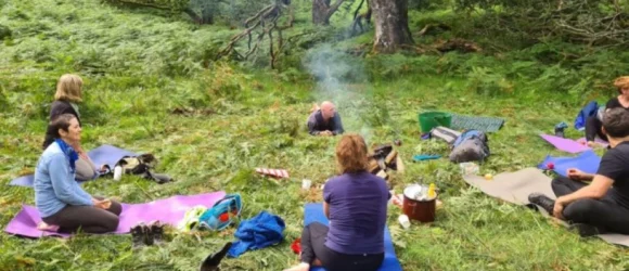 A group of people sitting on mats in a grassy forest clearing, surrounded by trees and foliage, with supplies and bags nearby.