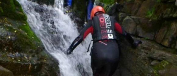 A person in an orange helmet and wetsuit climbing up a waterfall.