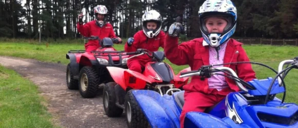 Children wearing helmets and red protective gear riding quad bikes on a dirt path.