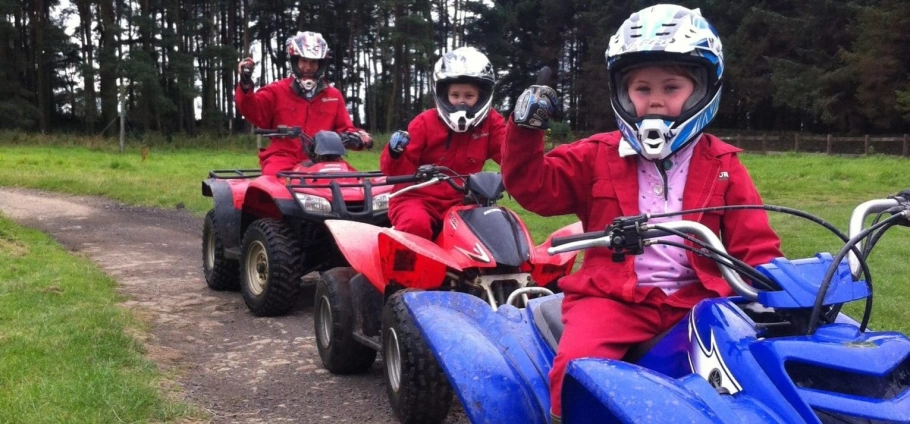 Children wearing helmets and red protective gear riding quad bikes on a dirt path.