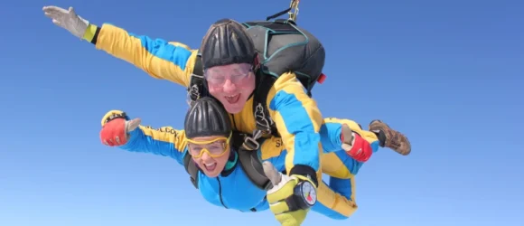 Two people tandem skydiving in colourful jumpsuits against a clear blue sky.