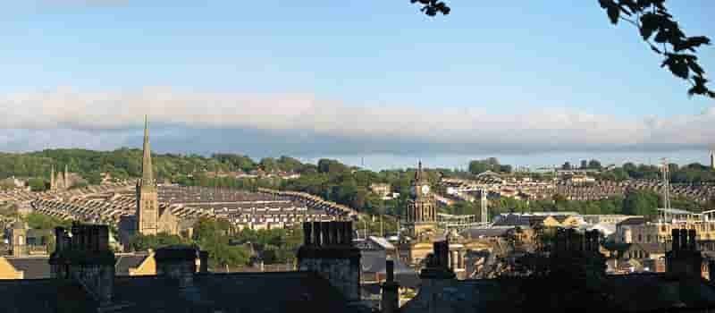 Historic Lancaster | A scenic view of a town with a prominent church spire and rows of terraced houses, surrounded by greenery under a partly cloudy sky.