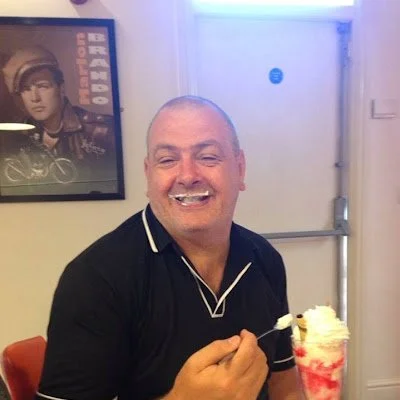 About 2 Smiling man with a moustache eating whipped cream from a dessert in a glass while sitting indoors with a framed picture of a man wearing a helmet on the wall behind him.