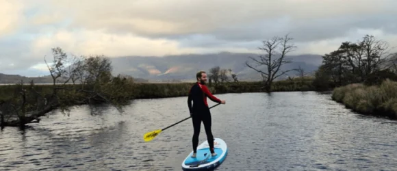 Person paddleboarding on calm water with a scenic, cloudy mountain range and trees in the background.