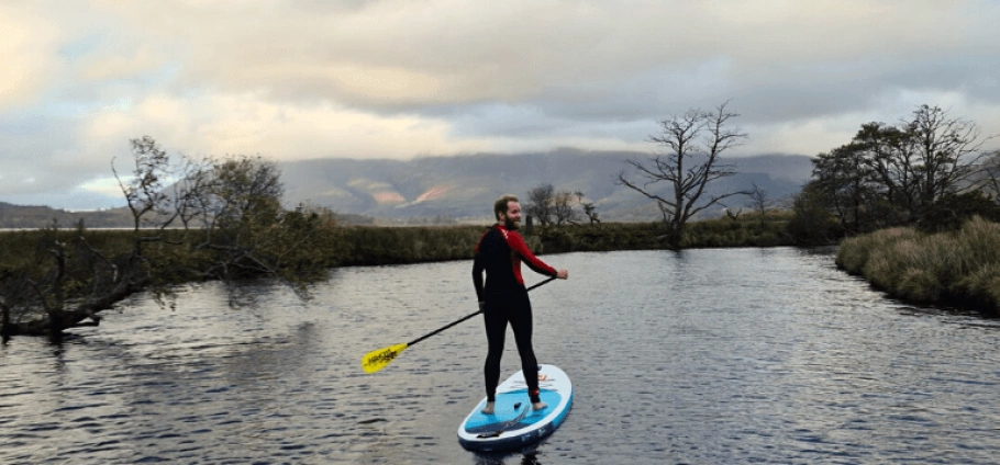 Person paddleboarding on calm water with a scenic, cloudy mountain range and trees in the background.