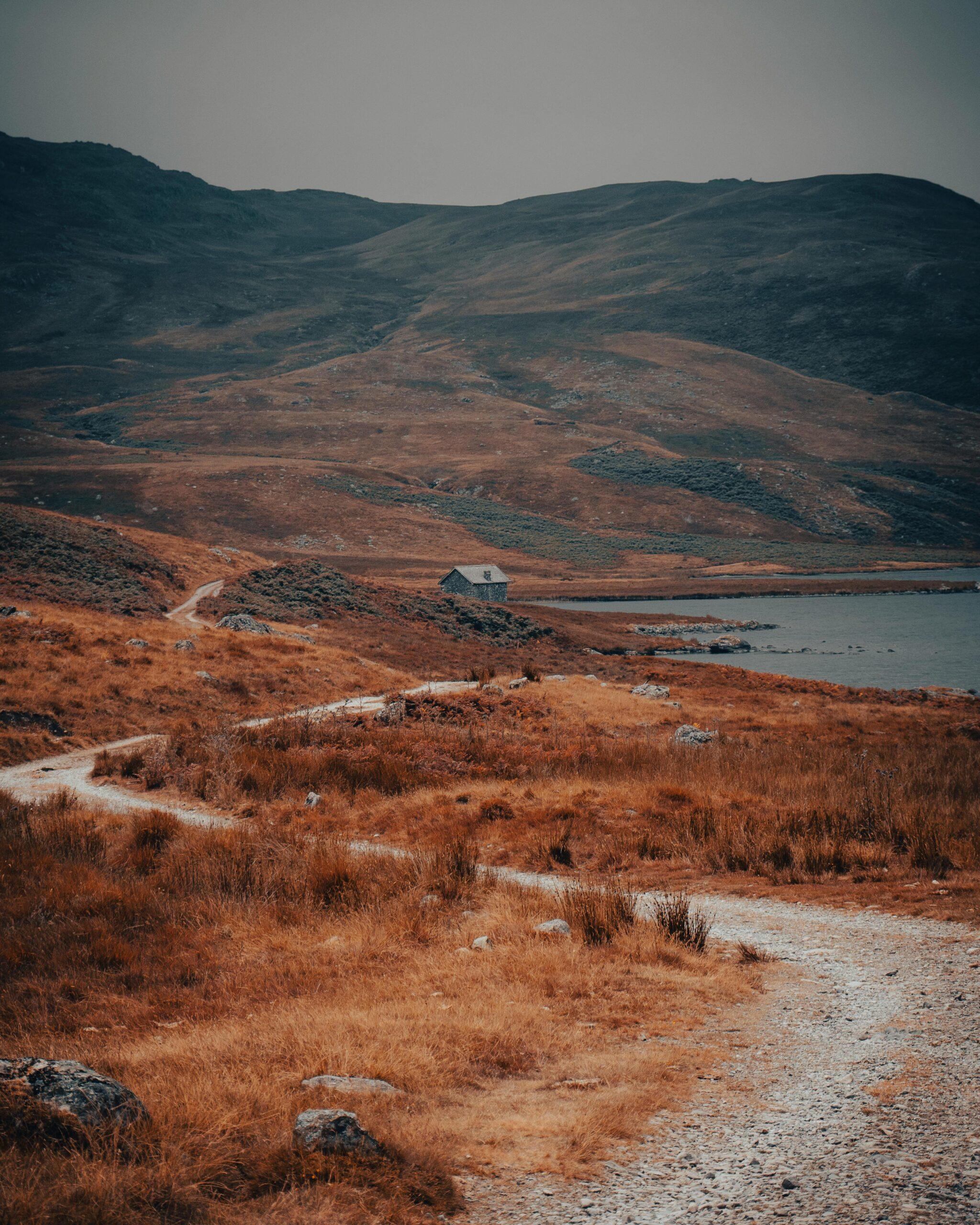 Ravenglass and Eskdale. A winding dirt path leading to a small house in a vast, rugged landscape with mountains in the background.