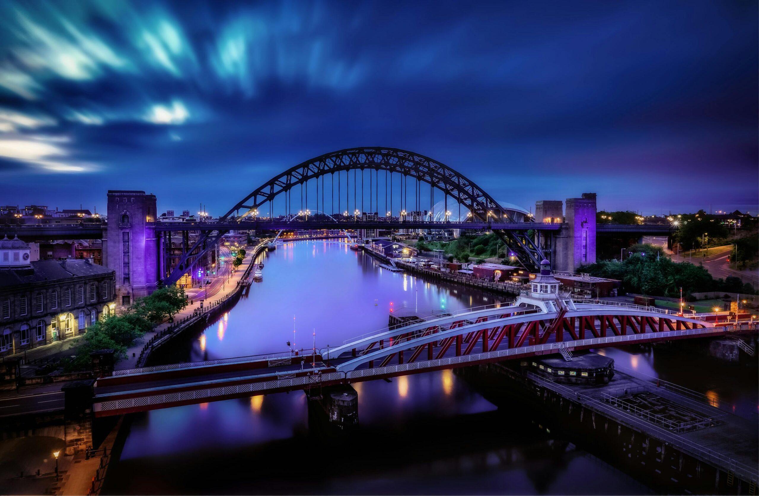 Night view of the Tyne Bridge over the River Tyne in Newcastle, illuminated with city lights reflecting on the water under a twilight sky.