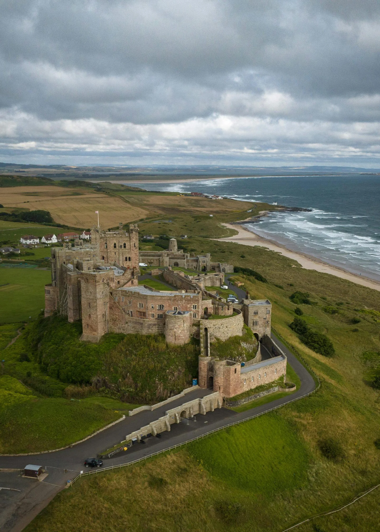 About 1 A large historic stone castle situated on a green hill near a coastline with a sandy beach, under a cloudy sky.
