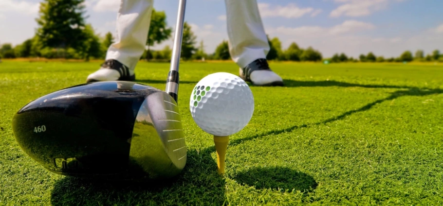 Golf club, ball, and tee on a golf course with a golfer's legs in the background.