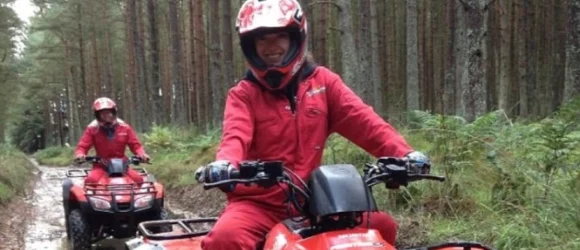 Two people wearing red jumpsuits and helmets riding quad bikes through a forest trail.