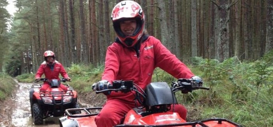 Two people wearing red jumpsuits and helmets riding quad bikes through a forest trail.