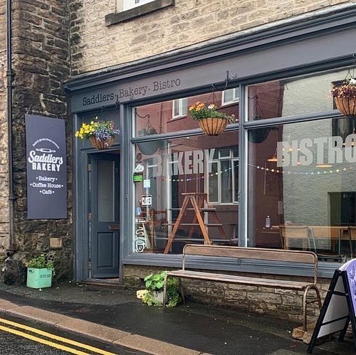 Exterior of Saddlers Bakery Bistro with a dark blue door and large windows displaying the words Bakery and Bistro, hanging flower baskets above, and a wooden bench in front on a wet pavement