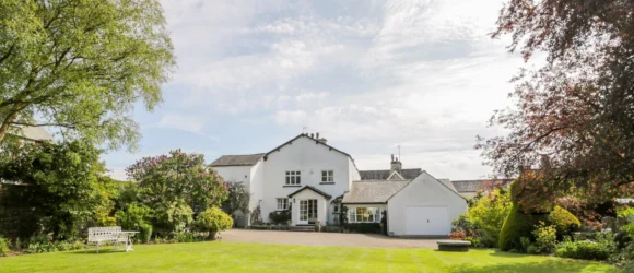 White house with a grey roof surrounded by a large, well-kept garden with green grass, trees, and bushes, featuring a white bench and a paved driveway.