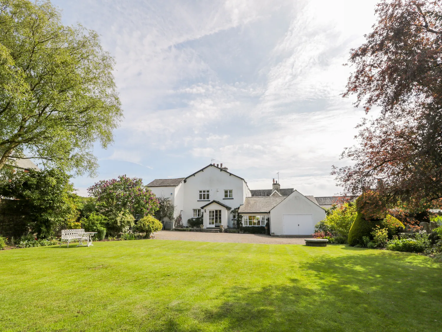 White house with a grey roof surrounded by a large, well-kept garden with green grass, trees, and bushes, featuring a white bench and a paved driveway.