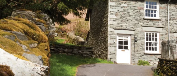 Stone cottage with white window frames and door, nestled next to a moss-covered stone wall and surrounded by greenery.