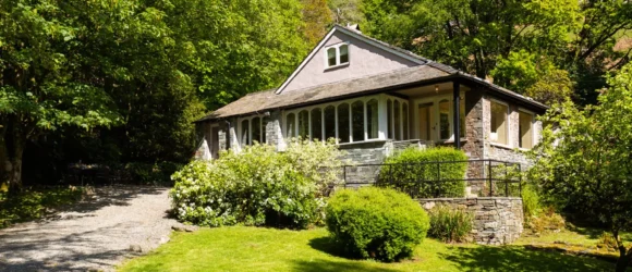 A charming cottage surrounded by lush greenery and trees, featuring a gravel driveway and a well-maintained garden.