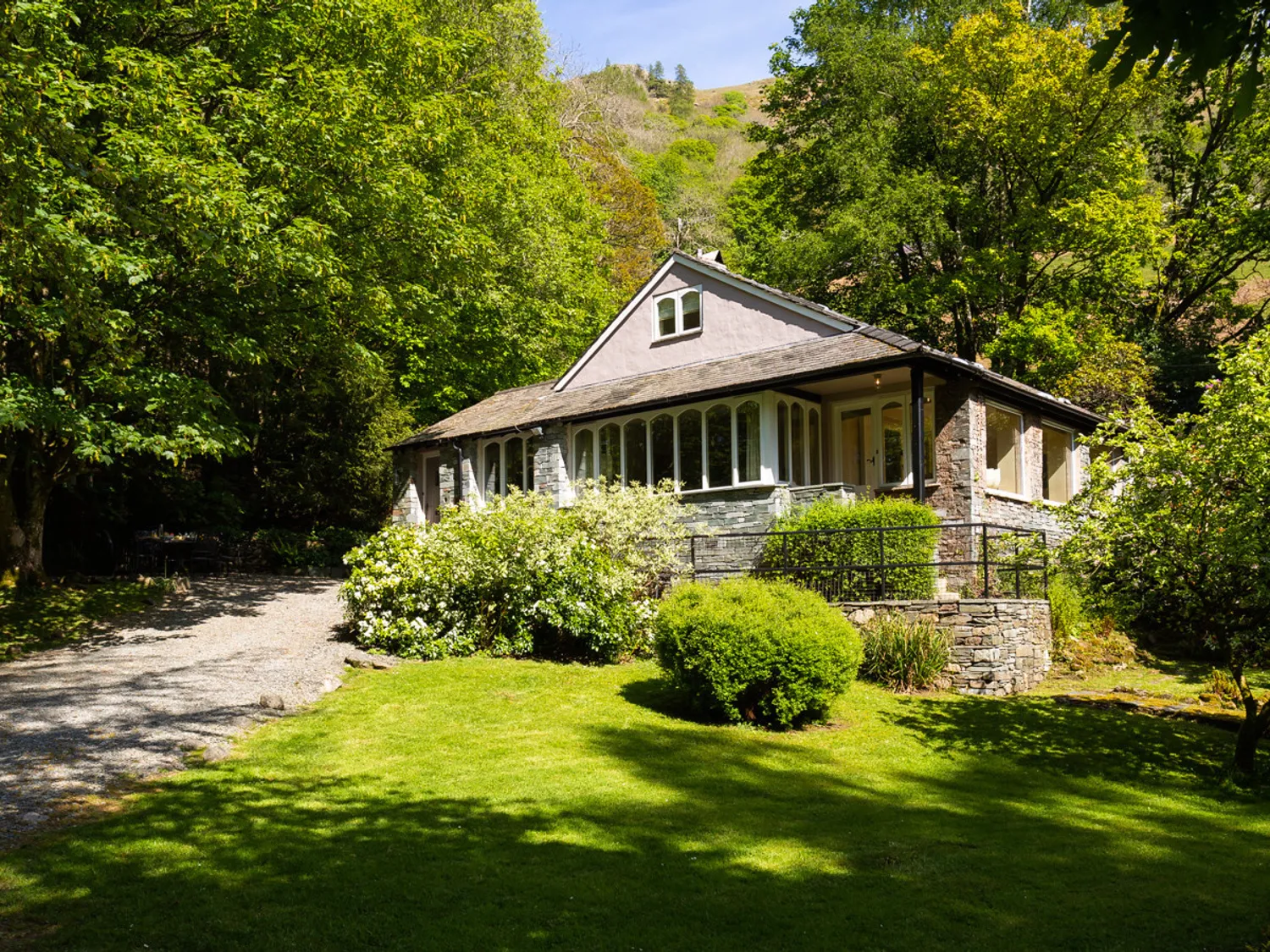 A charming cottage surrounded by lush greenery and trees, featuring a gravel driveway and a well-maintained garden.