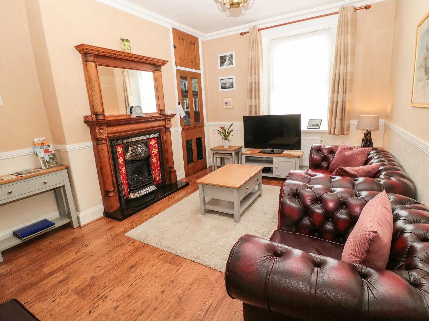 Cosy living room with dark leather sofas, a wooden coffee table, a flat-screen TV, a decorative fireplace with a mirror above, and light-coloured curtains.