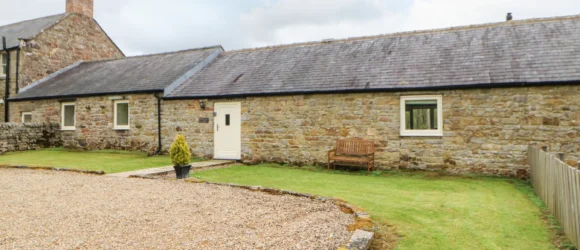 A stone cottage with a grey slate roof, white door, and gravel driveway, surrounded by a grassy lawn with a wooden bench and potted plant.