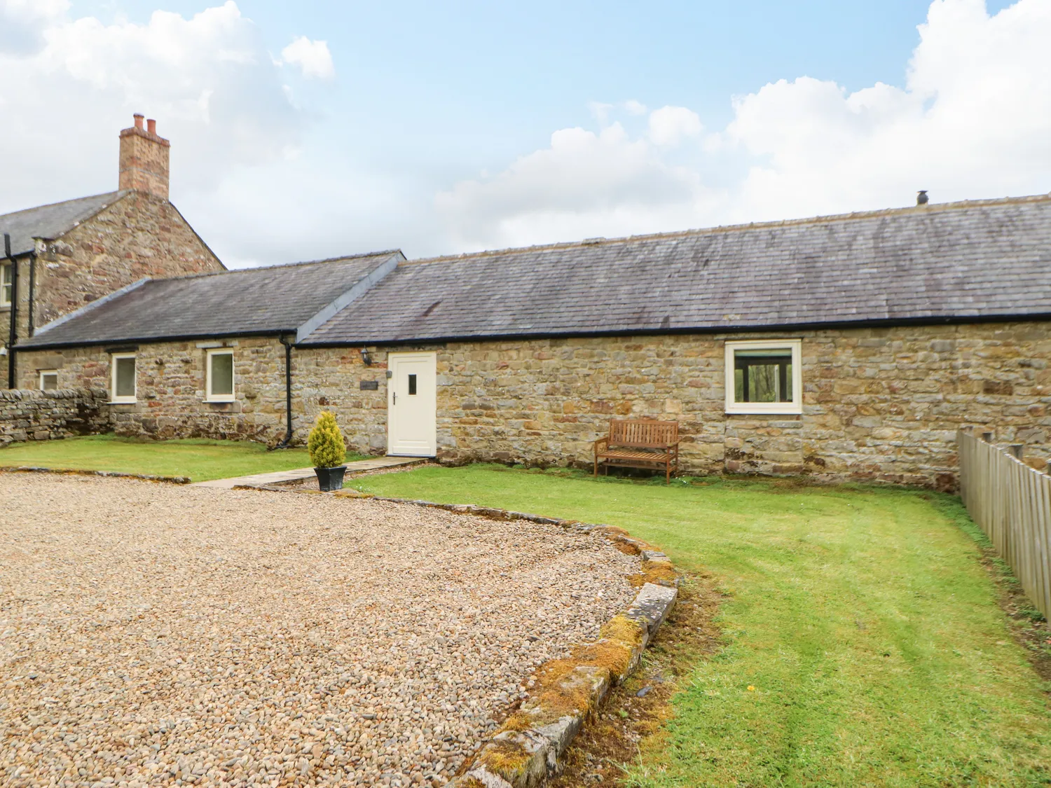 A stone cottage with a grey slate roof, white door, and gravel driveway, surrounded by a grassy lawn with a wooden bench and potted plant.