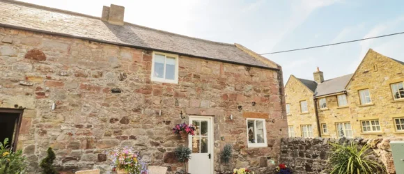 Stone cottage with a small, paved courtyard featuring a table and chairs, colourful flower planters, and a stone wall separating neighbouring buildings.