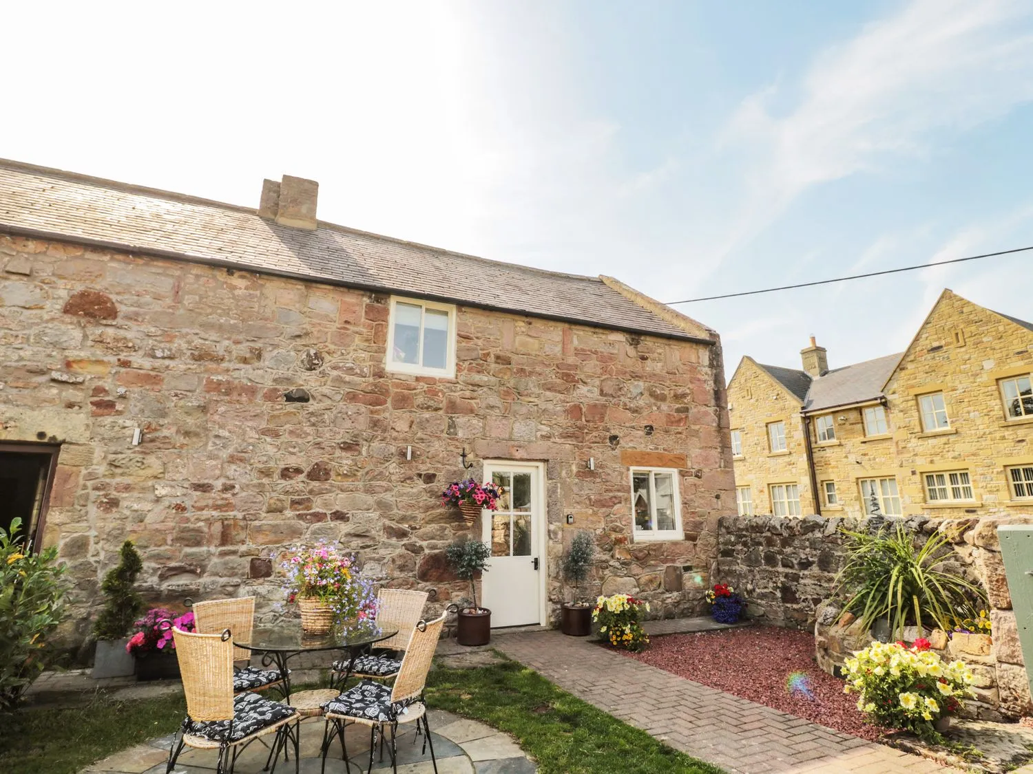 Stone cottage with a small, paved courtyard featuring a table and chairs, colourful flower planters, and a stone wall separating neighbouring buildings.