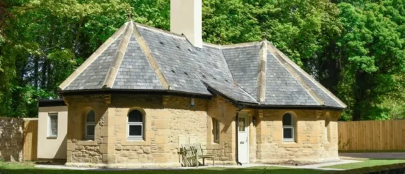 Octagonal stone cottage with a grey slate roof, set in a lush green wooded area.