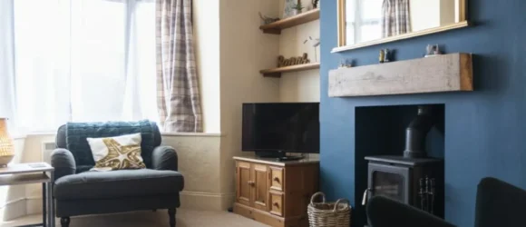 Cosy living room with a blue feature wall, wood-burning stove, wooden mantel, large mirror, armchair by the window, TV on a wooden stand, and plaid curtains.