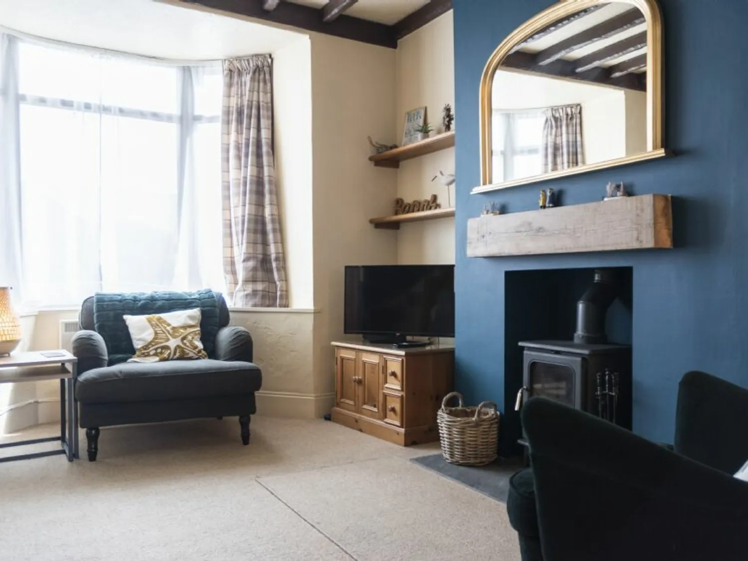 Cosy living room with a blue feature wall, wood-burning stove, wooden mantel, large mirror, armchair by the window, TV on a wooden stand, and plaid curtains.