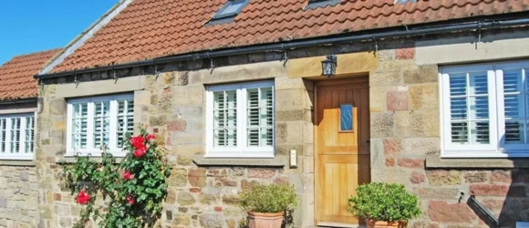 Stone cottage with a red tile roof, white-framed windows, and wooden front door, flanked by potted plants and climbing roses.