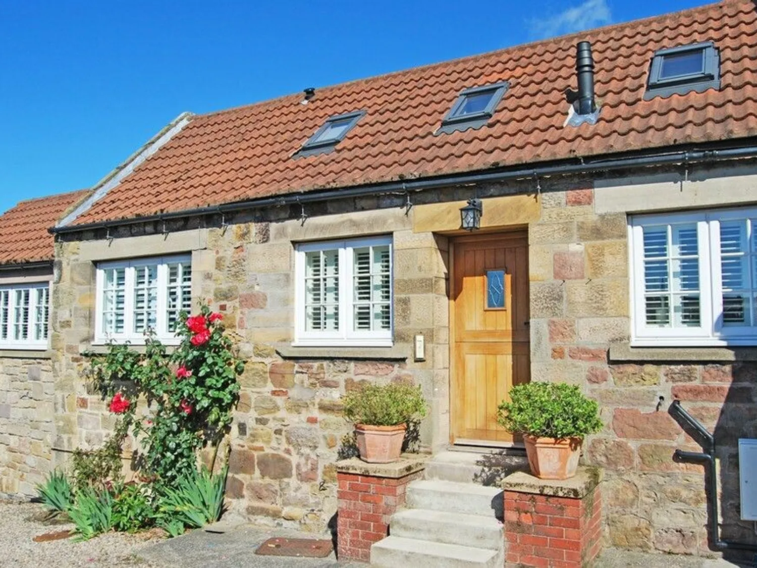 Stone cottage with a red tile roof, white-framed windows, and wooden front door, flanked by potted plants and climbing roses.