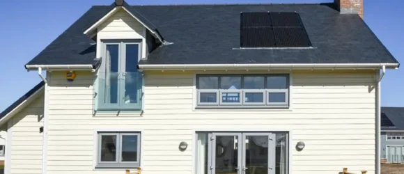 A modern two-story house with light-colored siding, a dark roof featuring solar panels, a small balcony with glass railing, large windows, and a wooden deck at the rear.