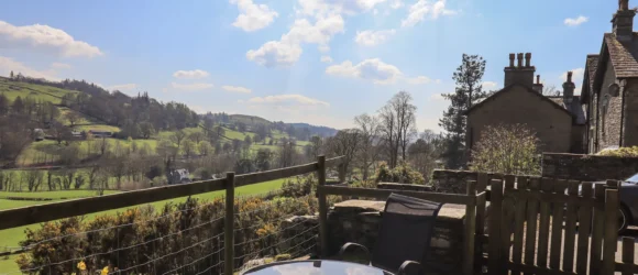 Patio area with a small table and chairs overlooking a scenic countryside with rolling green hills, trees, and scattered houses under a blue sky with scattered clouds.