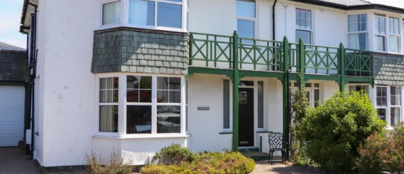 A semi-detached two-storey house with white walls and green trim, featuring bay windows and front garden with a black picket fence.