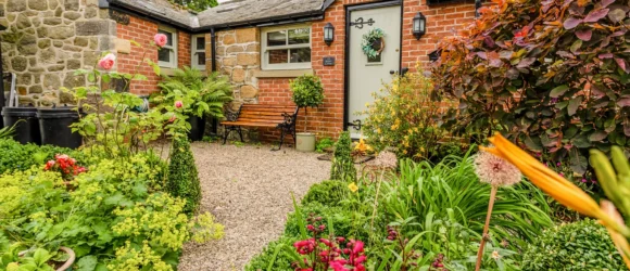 A quaint stone and brick house with a gable roof, surrounded by a lush, colourful garden featuring a variety of flowers and plants. A gravel path leads to the front door.